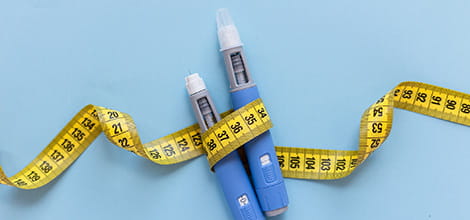 Two injectors dosing pens for subcutaneous injection of antidiabetic medication or anti-obesity medication hovering over a blue background. Yellow measuring tape around the injectors. Credit: iStock, Alones Creative.
